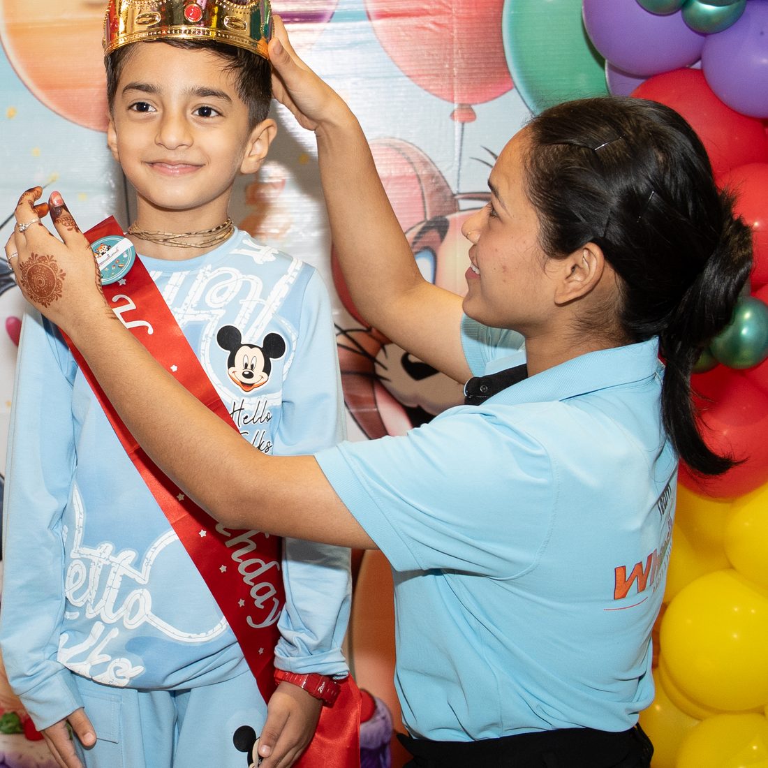 A WhimsiLand host crowning the birthday boy, sash on, balloons behind